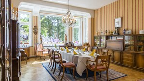 A dining room with a large table set for six, surrounded by wooden chairs, a chandelier, and a view of a garden through large windows. - Gästehaus Musmann (Hann. Muenden)