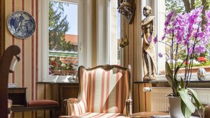 A room with a striped armchair, a wooden table, a potted plant, and a window with a view of trees and a building. - Gästehaus Musmann (Hann. Muenden)