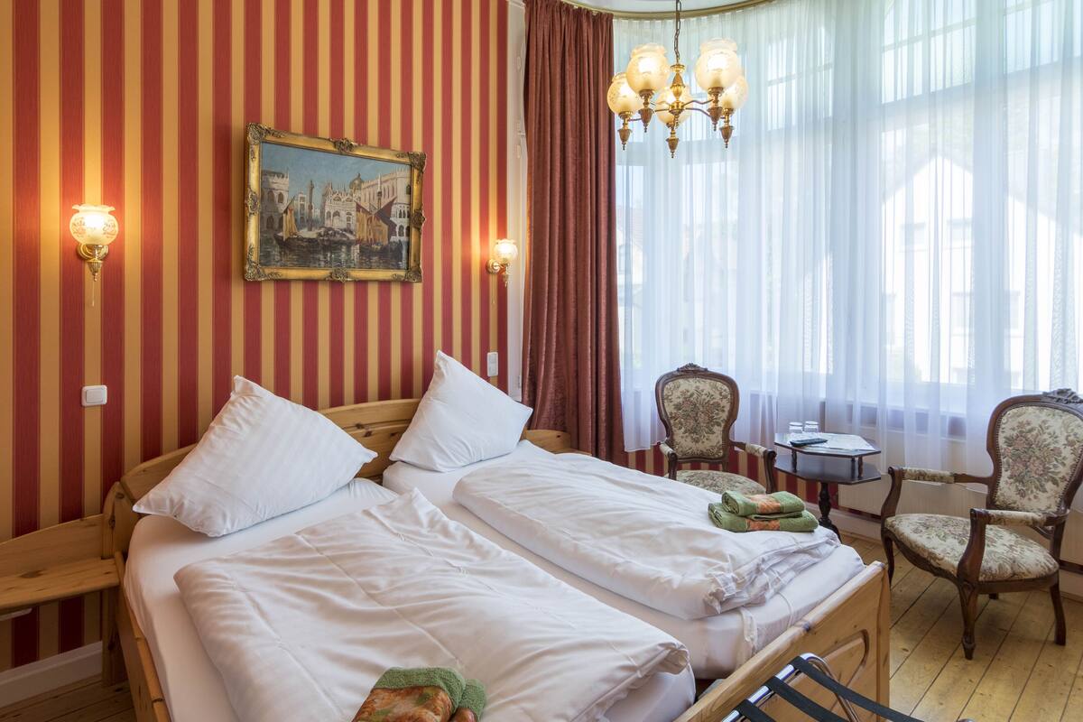 A hotel room with striped wallpaper, a bed with white linens, a painting of a cityscape, a chandelier, and a window with sheer curtains.