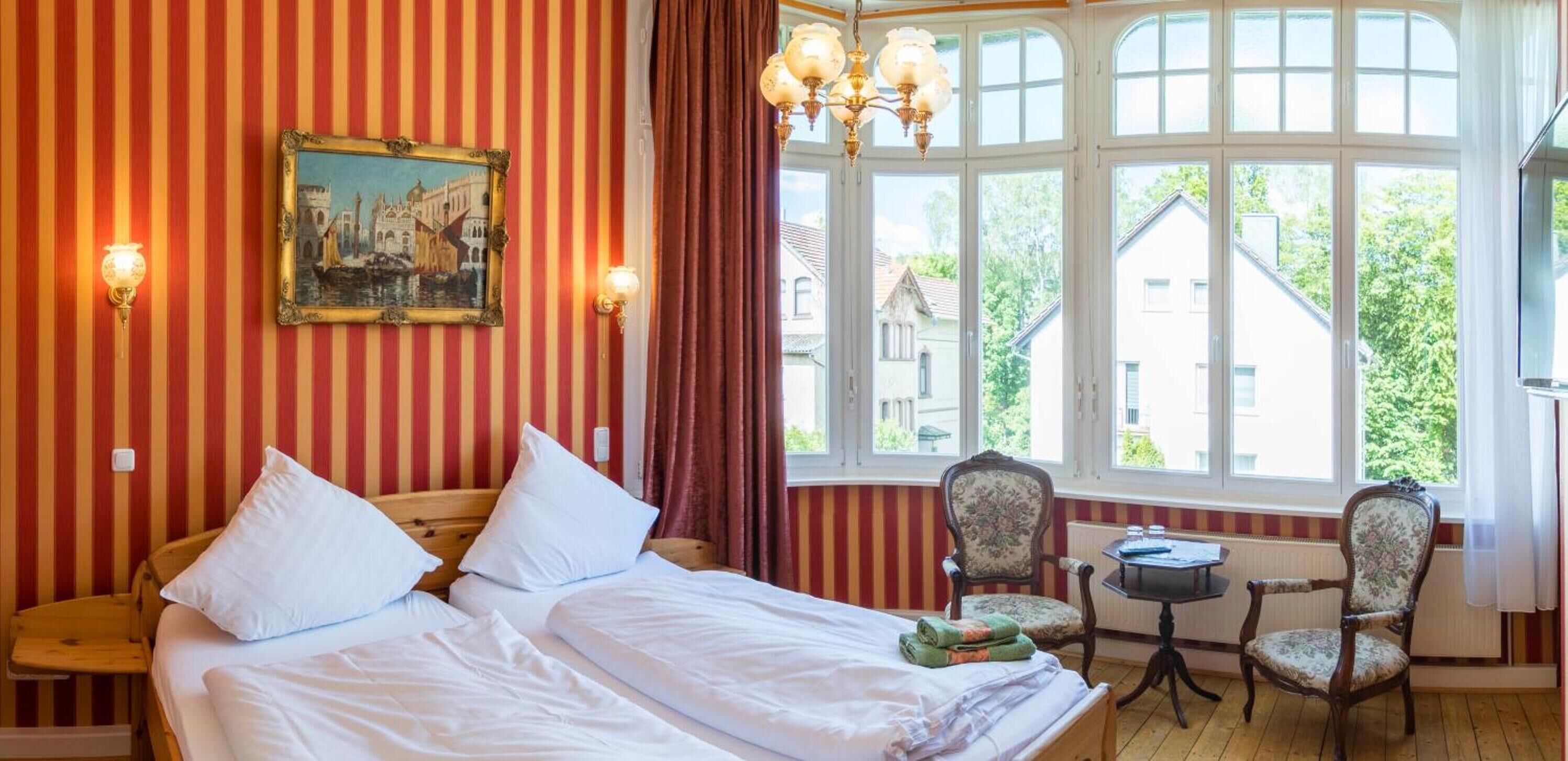A hotel room with a bed, two chairs, a table, a painting, and a window with a view of buildings.