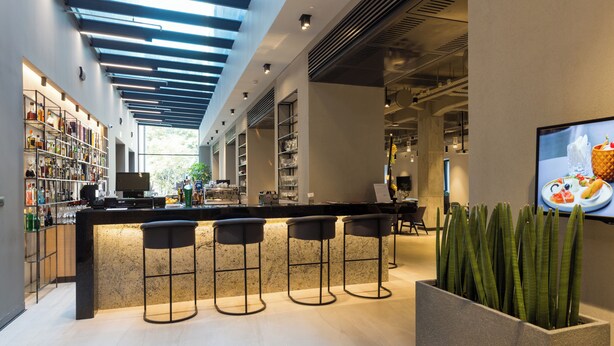Uma área de bar moderna com um balcão extenso, três banquetas e uma tela grande exibindo comida.