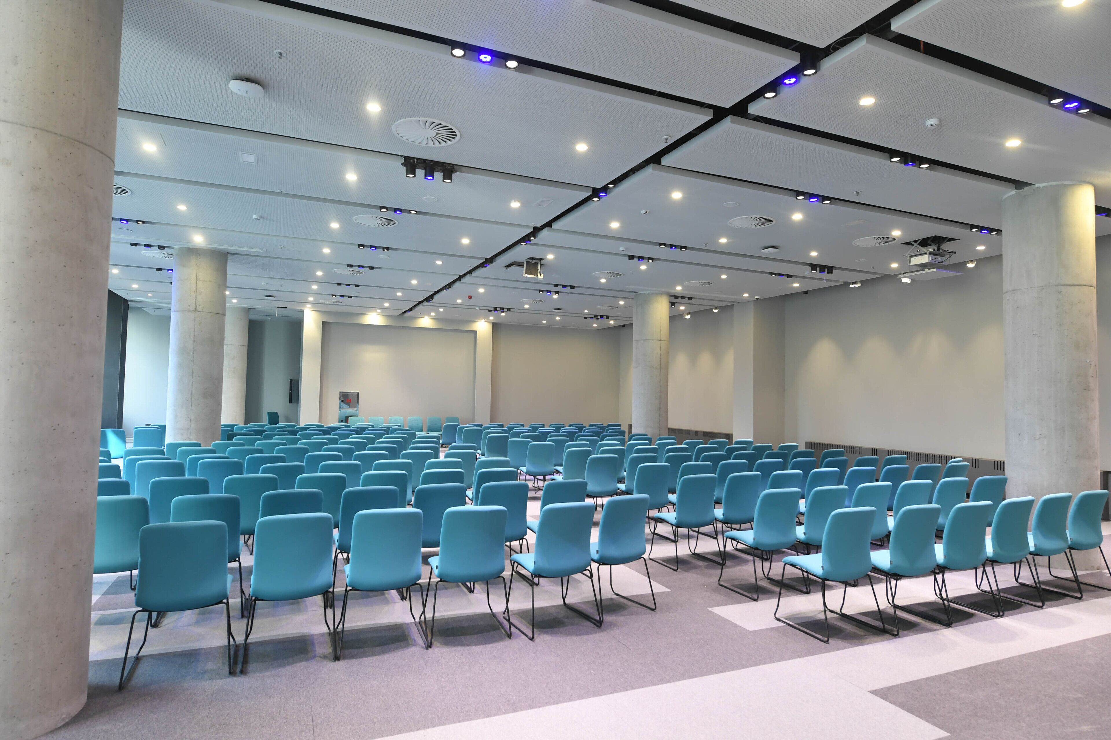 Un salón de conferencias con filas de sillas azules, paredes blancas e iluminación empotrada y rejillas de ventilación en el techo.