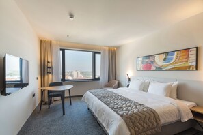 A hotel room with a bed, a desk, a chair, a TV, a window with curtains, and a painting on the wall. - Hotel Mona Plaza (Belgrade)