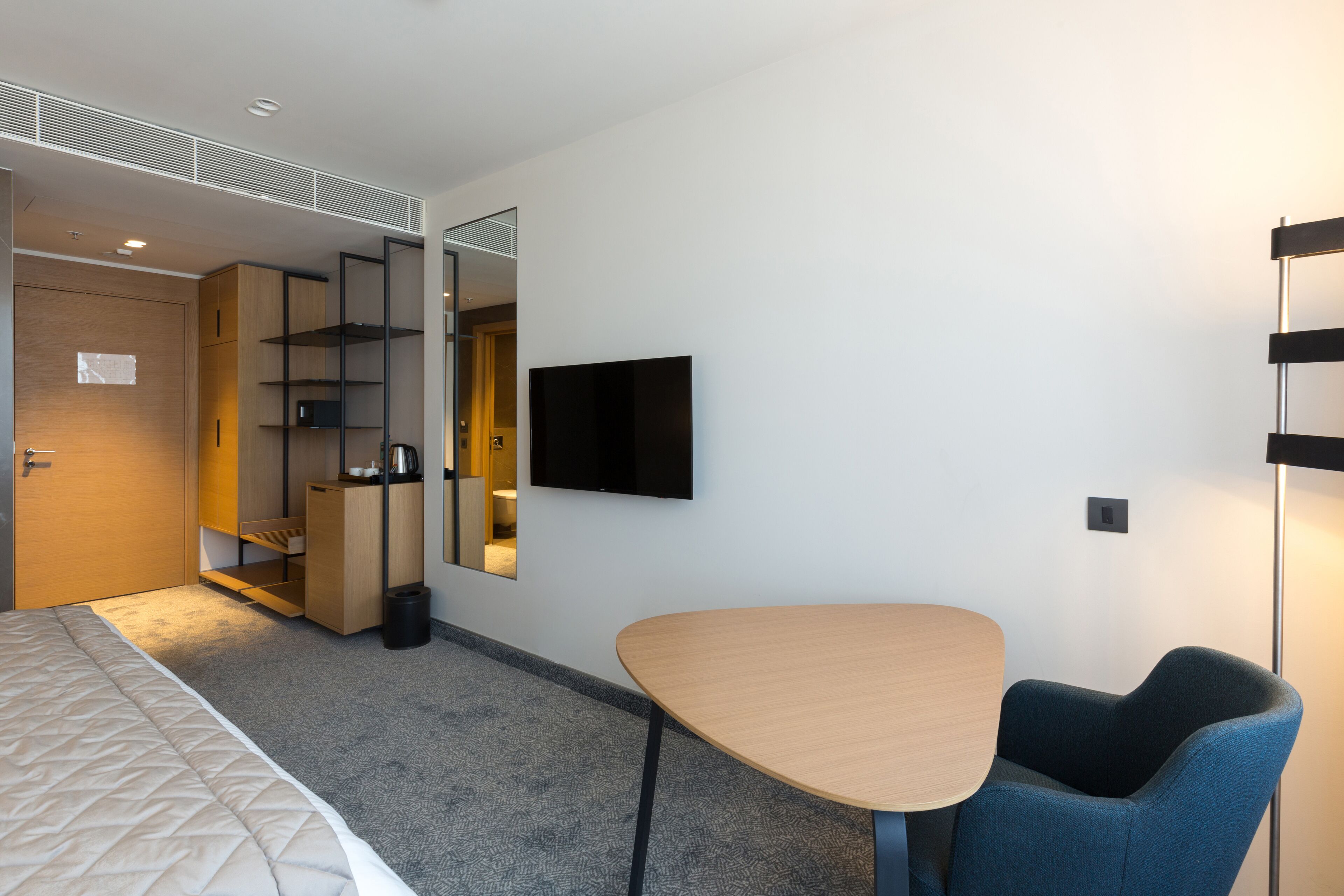 A modern hotel room with a bed, a desk, a chair, a TV, and built-in shelving.
