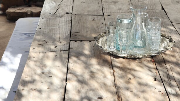 Una mesa de madera rústica con un jarro de vidrio y vasos sobre una bandeja, una escalera de piedra y un gato sentado en los escalones.