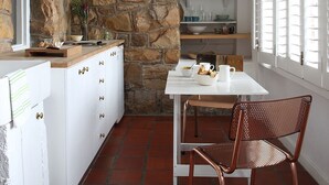 Una cocina con armarios blancos, pared de piedra y zona de comedor con mesa y sillas.