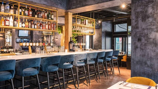 Eine Bar mit mehreren Regalen voller Spirituosenflaschen, einer Theke mit Zapfhähnen und verschiedenen blauen Barhocker.