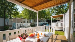 A covered outdoor dining area with a table set for breakfast, surrounded by white railings and a view of mobile homes. - Happy Camp in Camping Bella Italia (Peschiera del Garda)