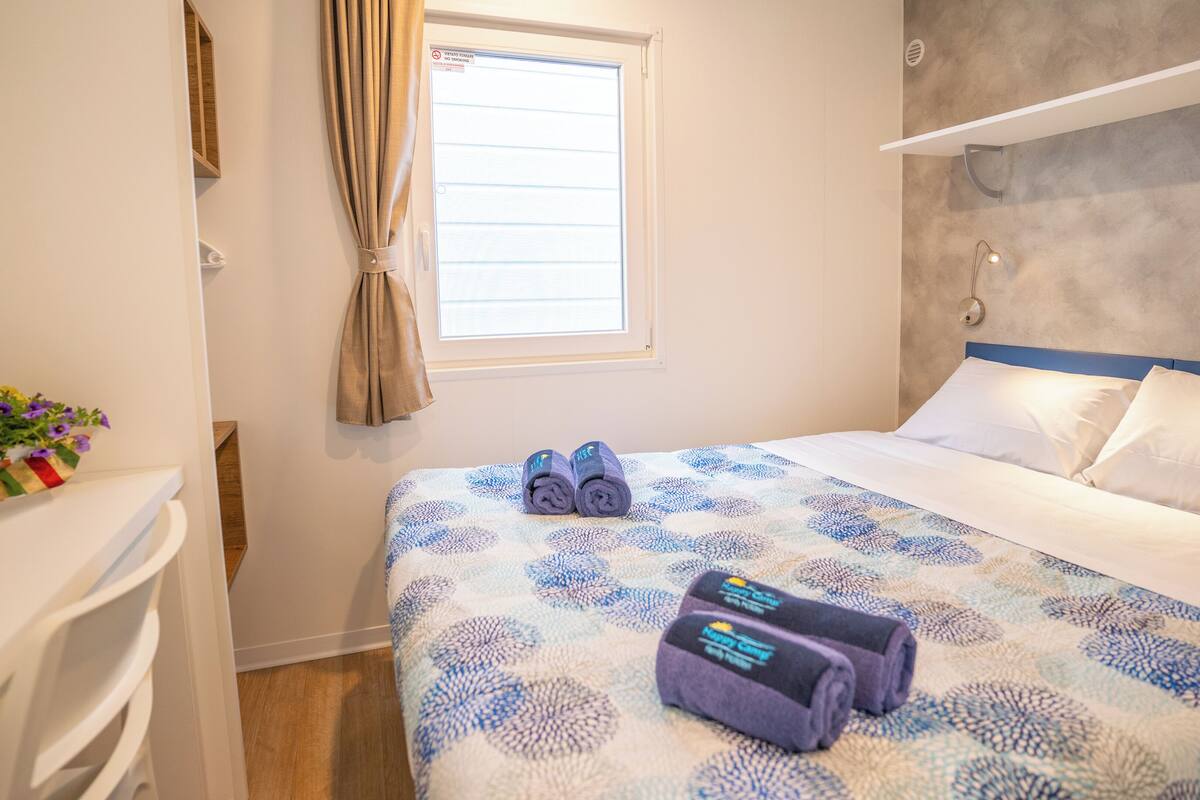 Una camera d'albergo con un letto, una finestra con tende, un comodino con un vaso di fiori e una testiera con una lampada.