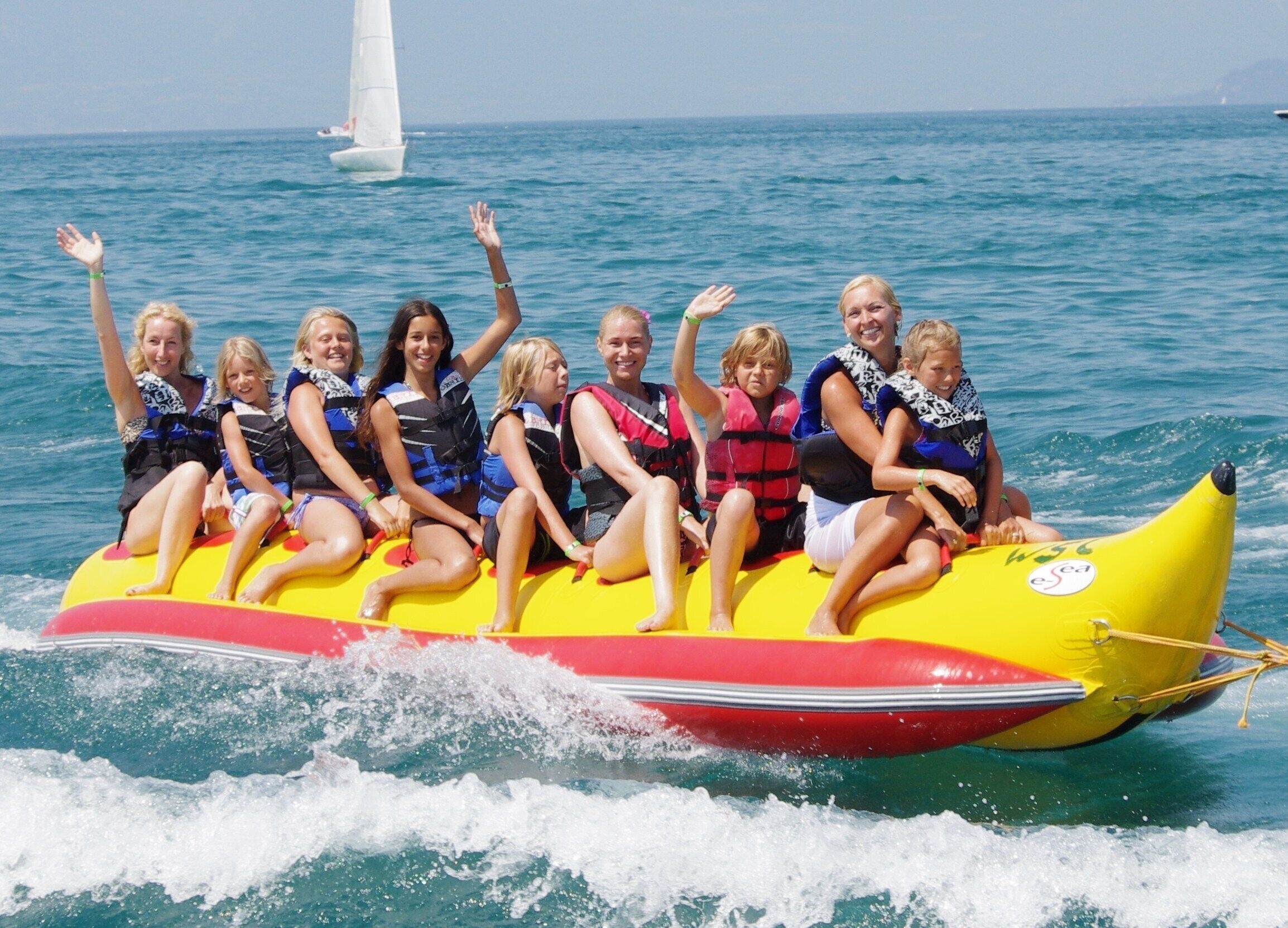 Eine Gruppe von Menschen auf einem bananenförmigen Floß im Meer, im Hintergrund ein Segelboot.