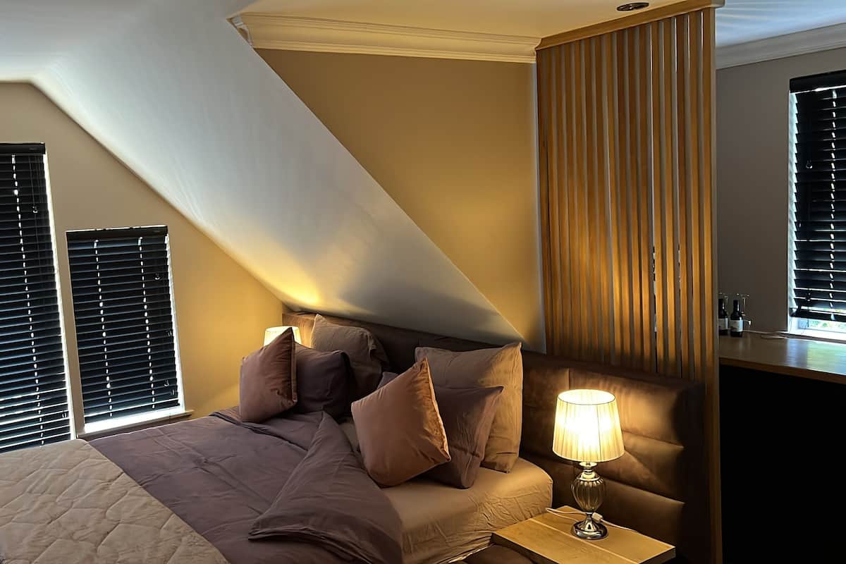 Une chambre à coucher avec un lit, des oreillers, une table de chevet, une lampe et une fenêtre avec des stores.