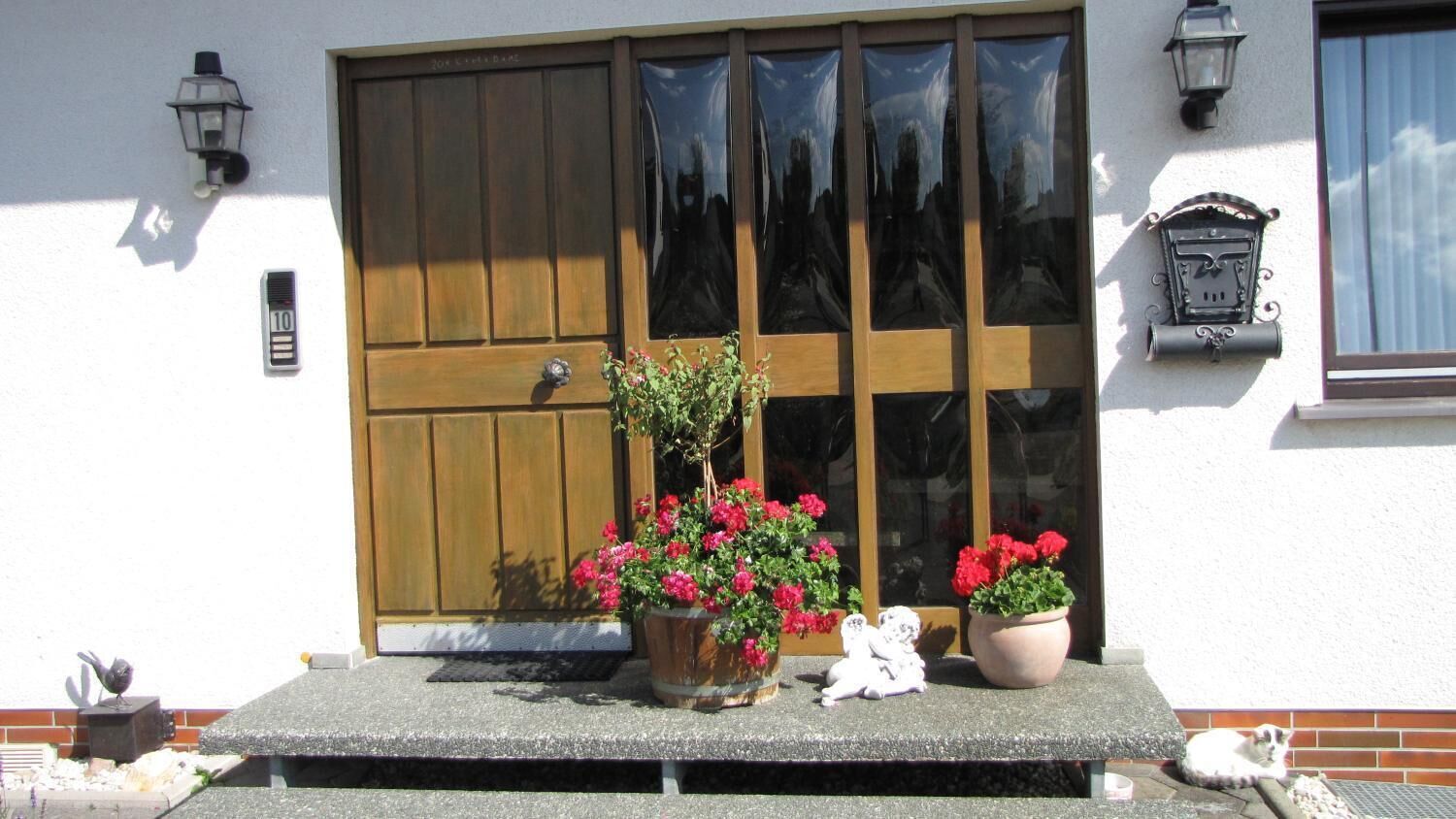 A building entrance with a wooden door, two potted plants with red flowers, a small white figurine, and a mailbox.