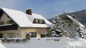 Una casa cubierta de nieve con techo inclinado, una chimenea y un ventanal con cortina.