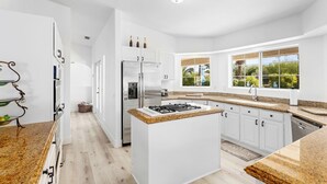 A modern kitchen with white cabinetry, granite countertops, a central island, and a view of greenery through large windows. - Palm View Ranch - 1 acre mini resort! (Palm Desert)