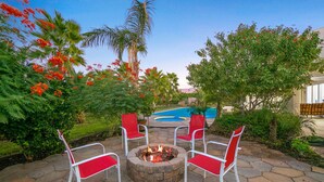 A patio area with a fire pit, four red and white chairs, and a swimming pool in the background. - Palm View Ranch - 1 acre mini resort! (Palm Desert)