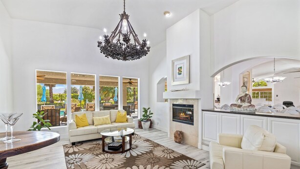 A spacious living room with a fireplace, a large chandelier, and a view of the outdoors through large windows. - Palm View Ranch - 1 acre mini resort! (Palm Desert)