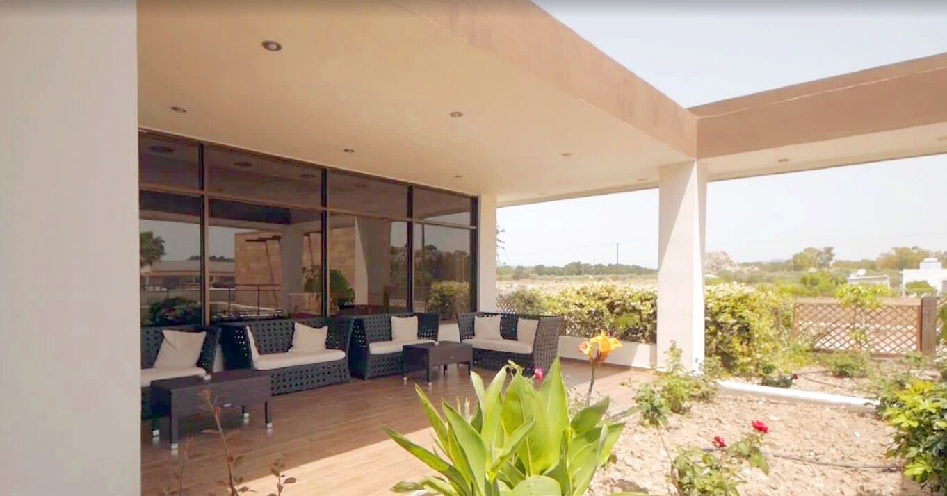 Uma área de estar moderna ao ar livre, com deck de madeira e paredes de vidro, com vista para um jardim paisagístico.