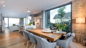 A modern kitchen with a large island, a dining area with a wooden table and grey chairs, and a large window offering a view of the outdoors. - "Heimathafen 2" vacation apartment with sauna, south-facing terrace & roof terrace by the water (Olpenitz)