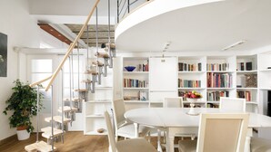 Ein geräumiger Raum mit einem weißen Esstisch, mehreren Stühlen, einer Wendeltreppe und einem Bücherregal voller Bücher.