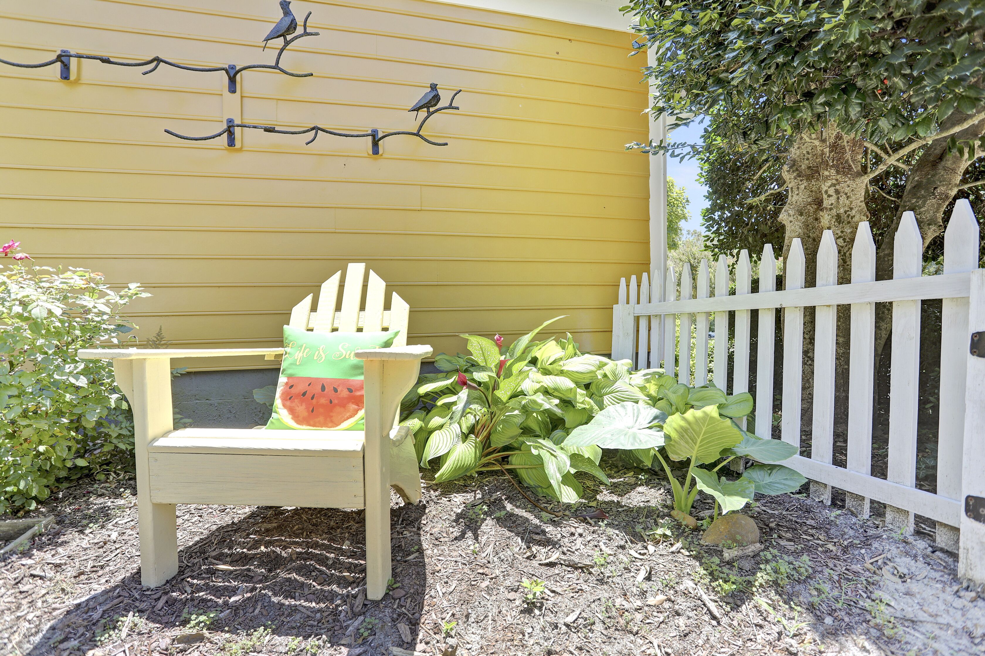 A garden area with a white picket fence, a bench with a watermelon cushion, and a yellow wall with birdhouses.