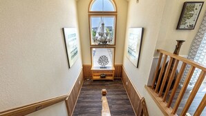Una escalera de madera con vistas al exterior, una pequeña mesa con una lámpara y obras de arte enmarcadas en las paredes.