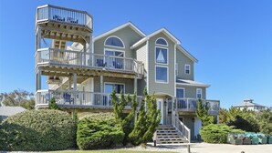 Una casa de varios pisos frente a la playa, con balcones, una escalera que lleva a la entrada y un césped bien cuidado.