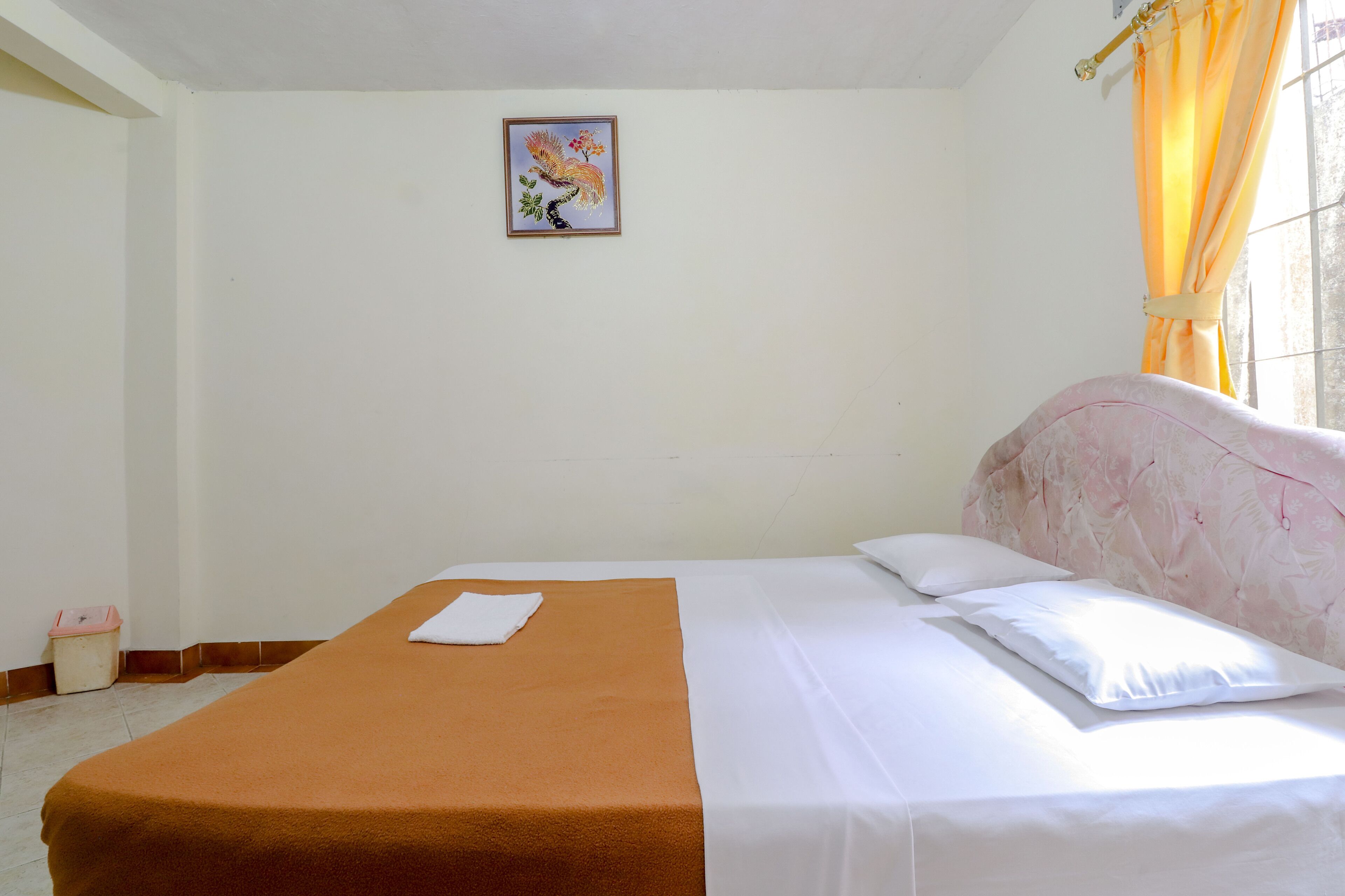 Una cama bien hecha con una colcha marrón y ropa de cama blanca, un cuadro en la pared y una ventana con cortinas.