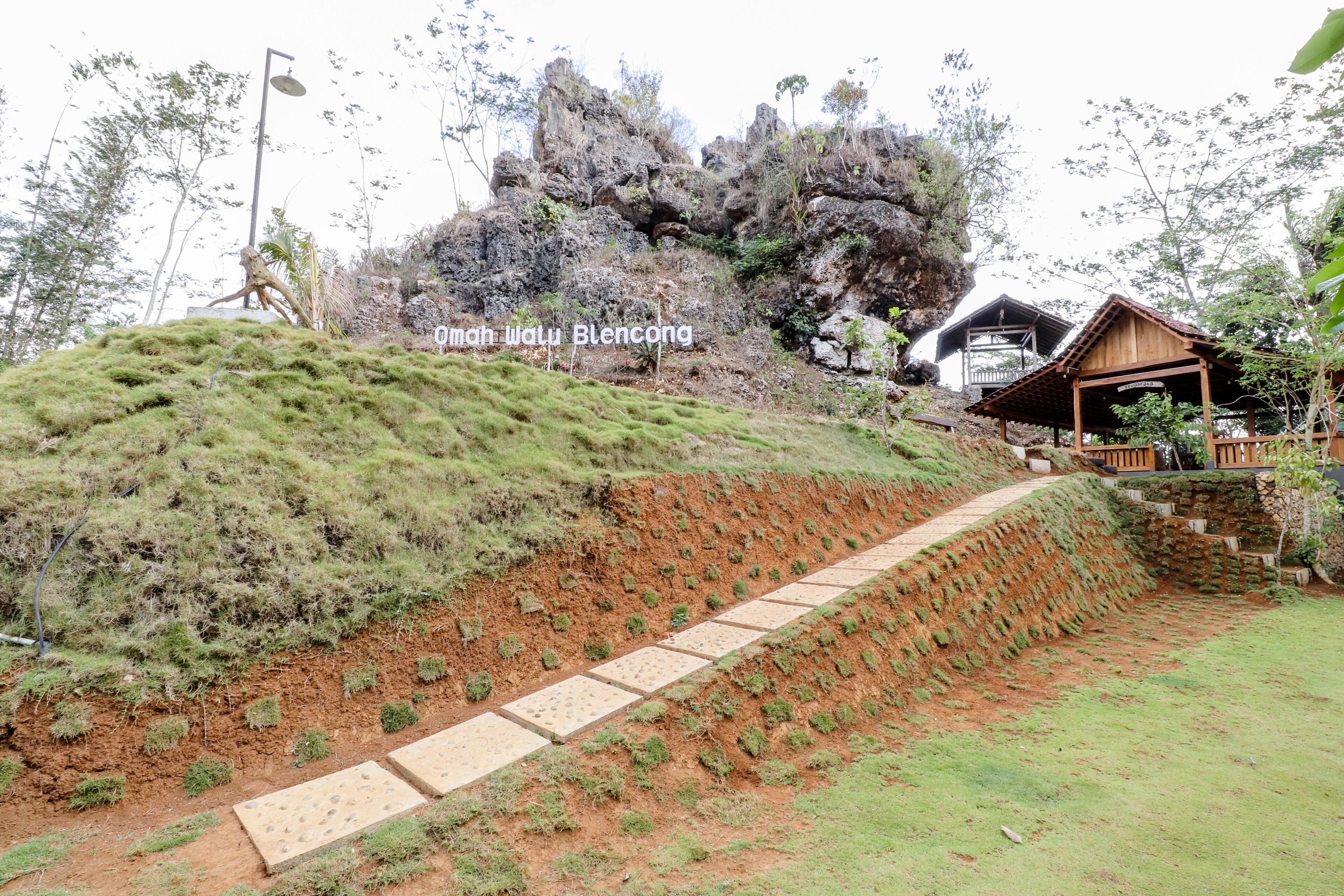 A terraced pathway leading to a wooden structure with a balcony, surrounded by greenery and a rocky backdrop.