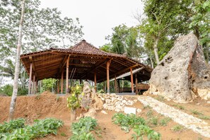 A rustic wooden pavilion with a tiled roof, surrounded by a dirt path and greenery, including a large rock formation and trees. - Omah Watu Blencong (Kulon Progo)