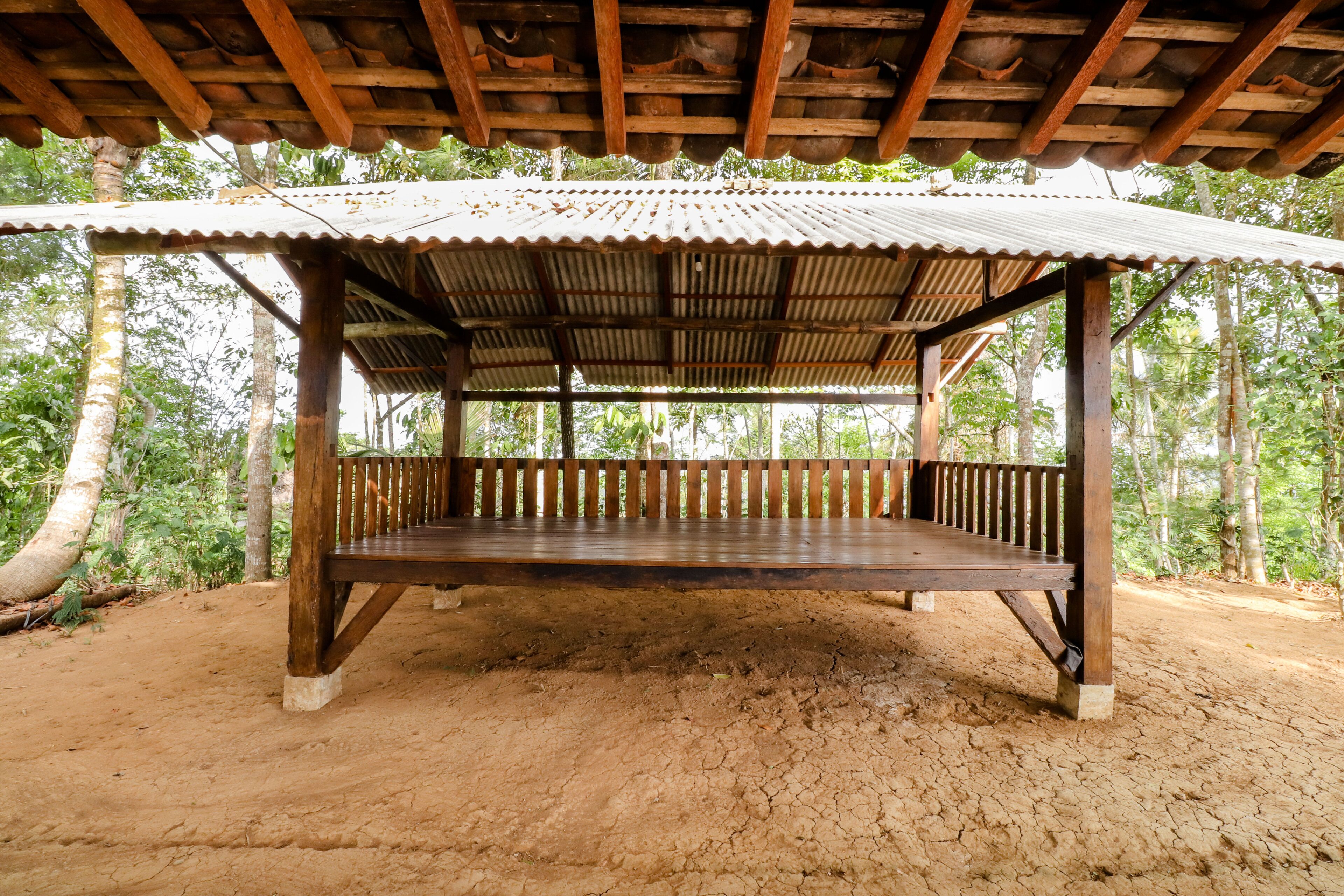 A wooden gazebo with a corrugated metal roof, supported by wooden pillars, set on sandy ground with trees in the background.