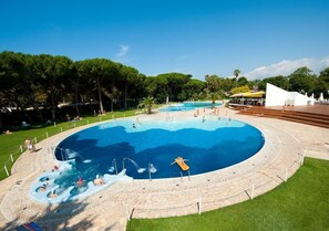 A large outdoor swimming pool with a circular design, surrounded by a stone deck and a grassy area. - Happy Camp in Camping Baia Domizia (Baia Domizia)