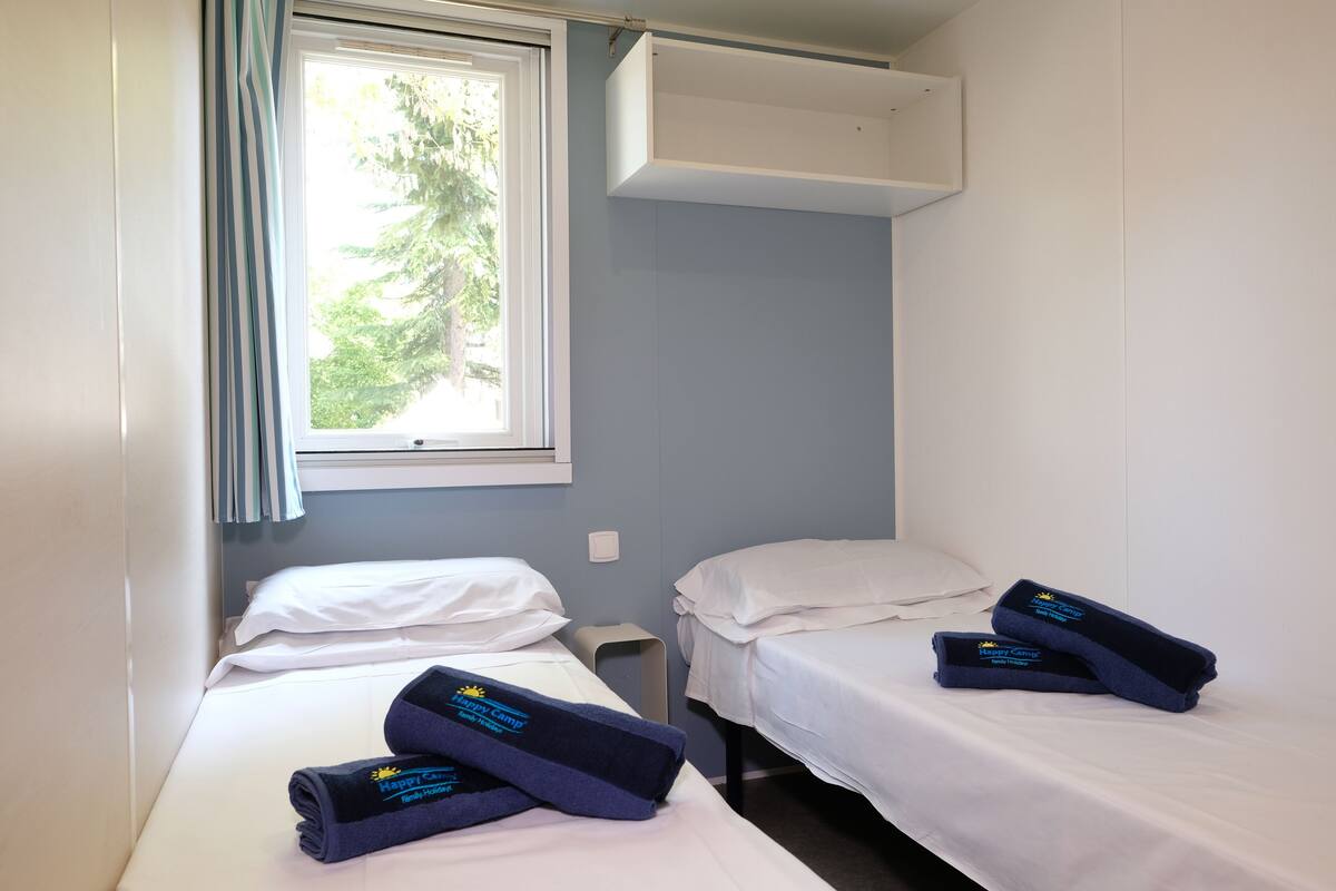 Ein Zimmer mit zwei Betten, jeweils mit weißer Matratze und Kissen, und einem Fenster mit Blick auf Bäume. Auf den Betten liegen Handtücher und über dem Fenster ein kleiner, weißer Schrank.