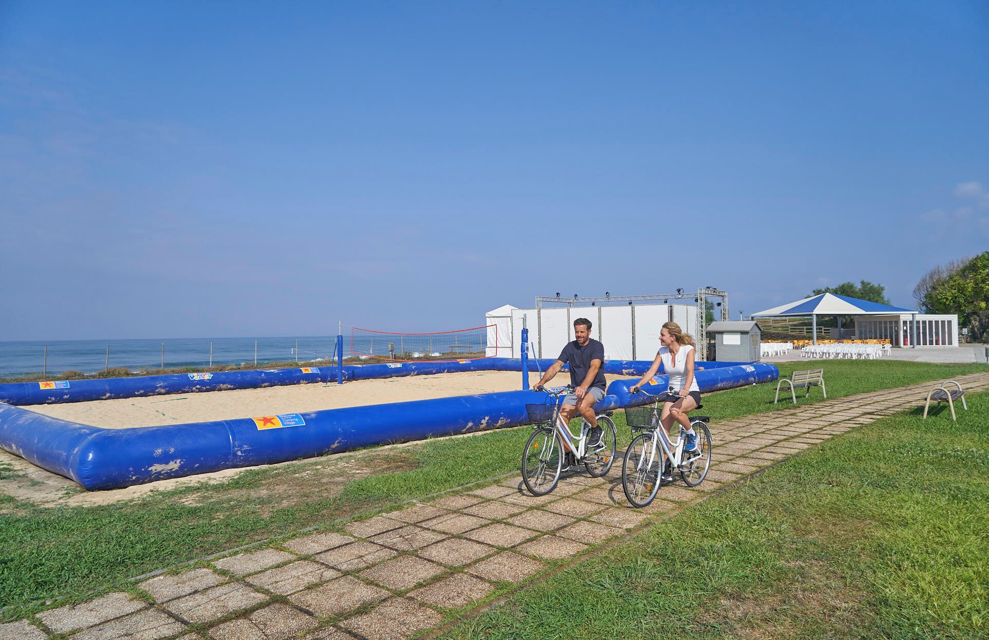 Un camino pavimentado con dos bicicletas, una barrera inflable azul y un edificio blanco con techo azul al fondo.