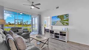 A modern living room with a large window, a ceiling fan, a flat-screen TV, and a wooden entertainment unit. - WATERLILY at Greenlinks - Updated Condo with Captivating Views! (Naples)