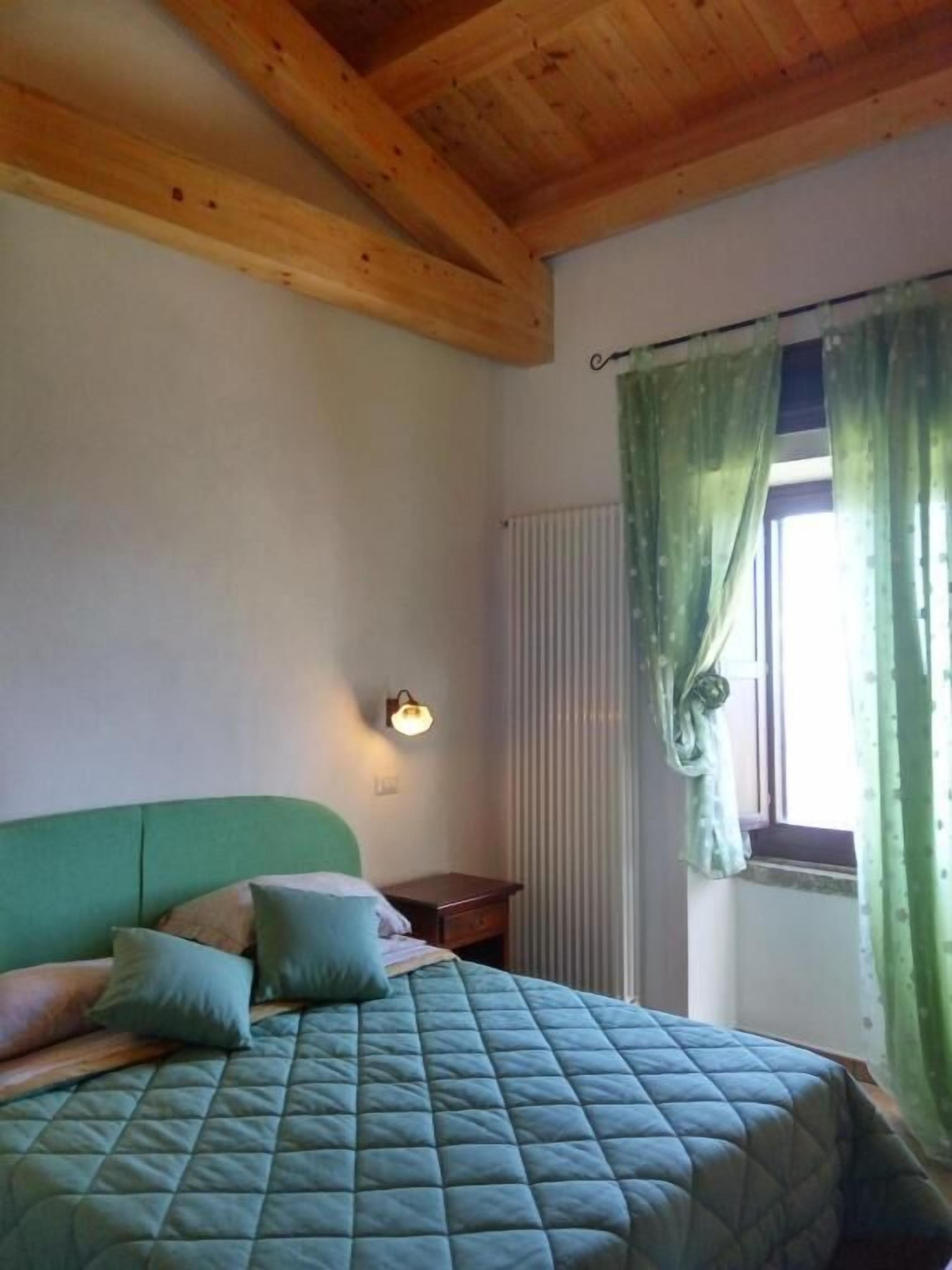 Un dormitorio con una cama verde, una mesita de noche, una lámpara de pared y una ventana con cortinas verdes.