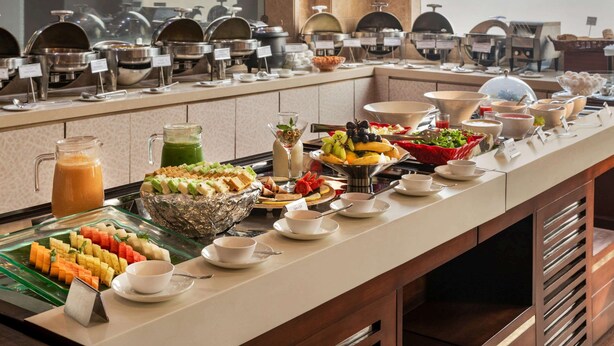 Un bufé con diversas opciones de comida y bebidas, incluyendo frutas, ensaladas y platos calientes, dispuestas en una larga barra.