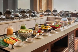 Un bufé con diversas opciones de comida y bebidas, incluyendo frutas, ensaladas y platos calientes, dispuestas en una larga barra.