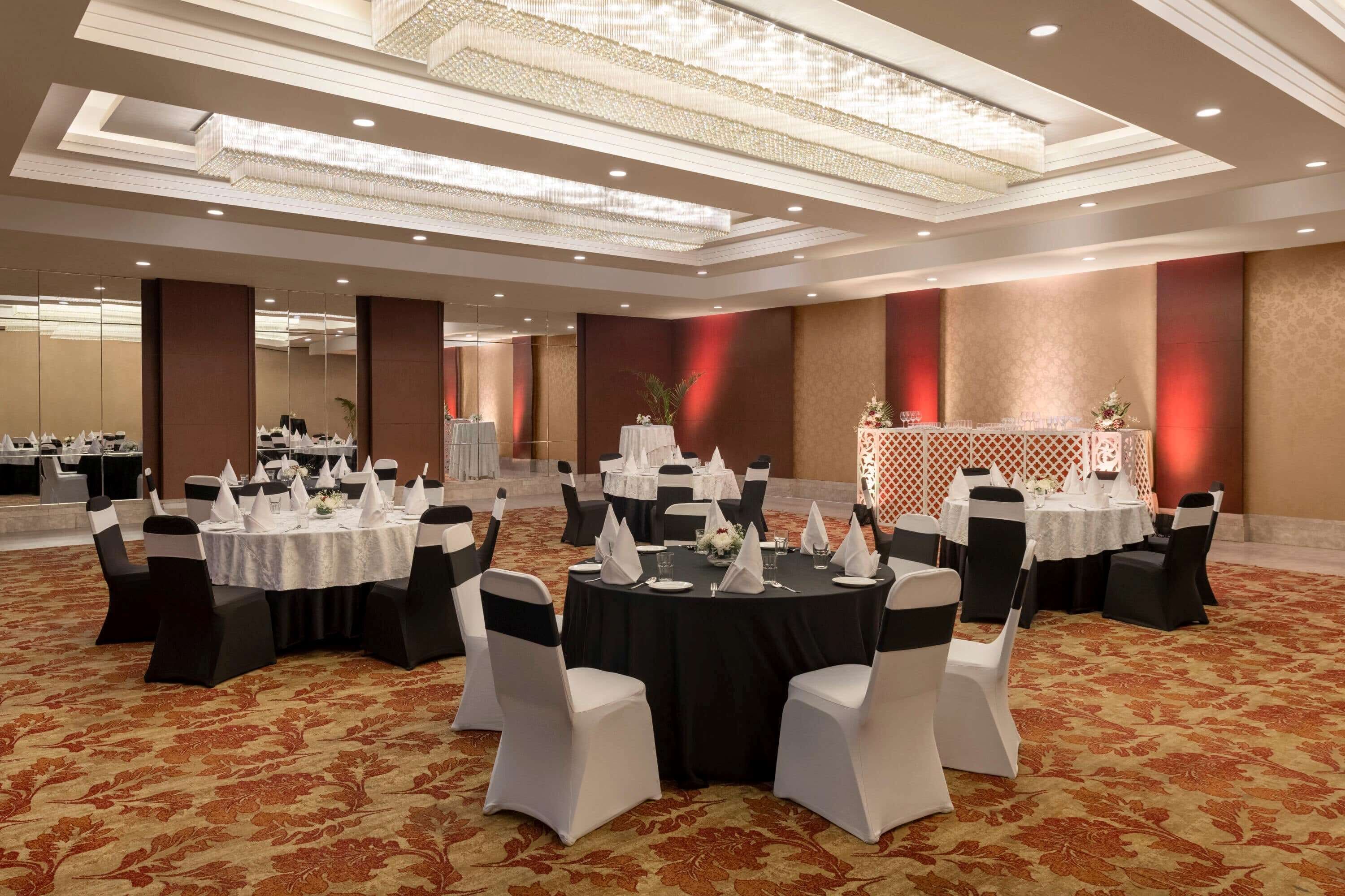 A banquet hall with round tables, black and white chairs, and a patterned carpet.