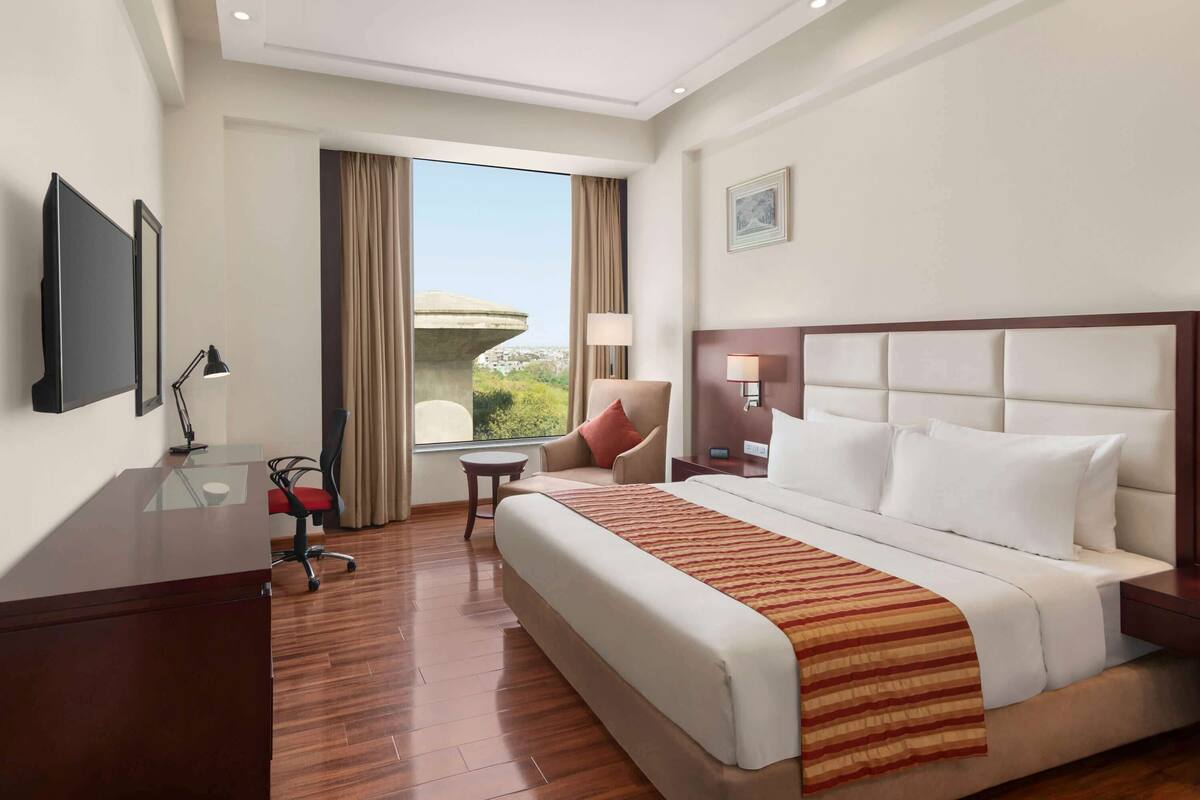 A hotel room with a large bed, a desk, a chair, a TV, and a window with a view of greenery.