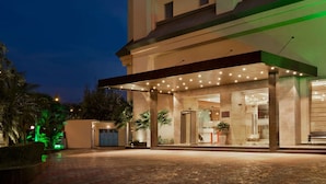 Entrada de hotel con zona exterior techada, palmeras y una fachada bien iluminada.