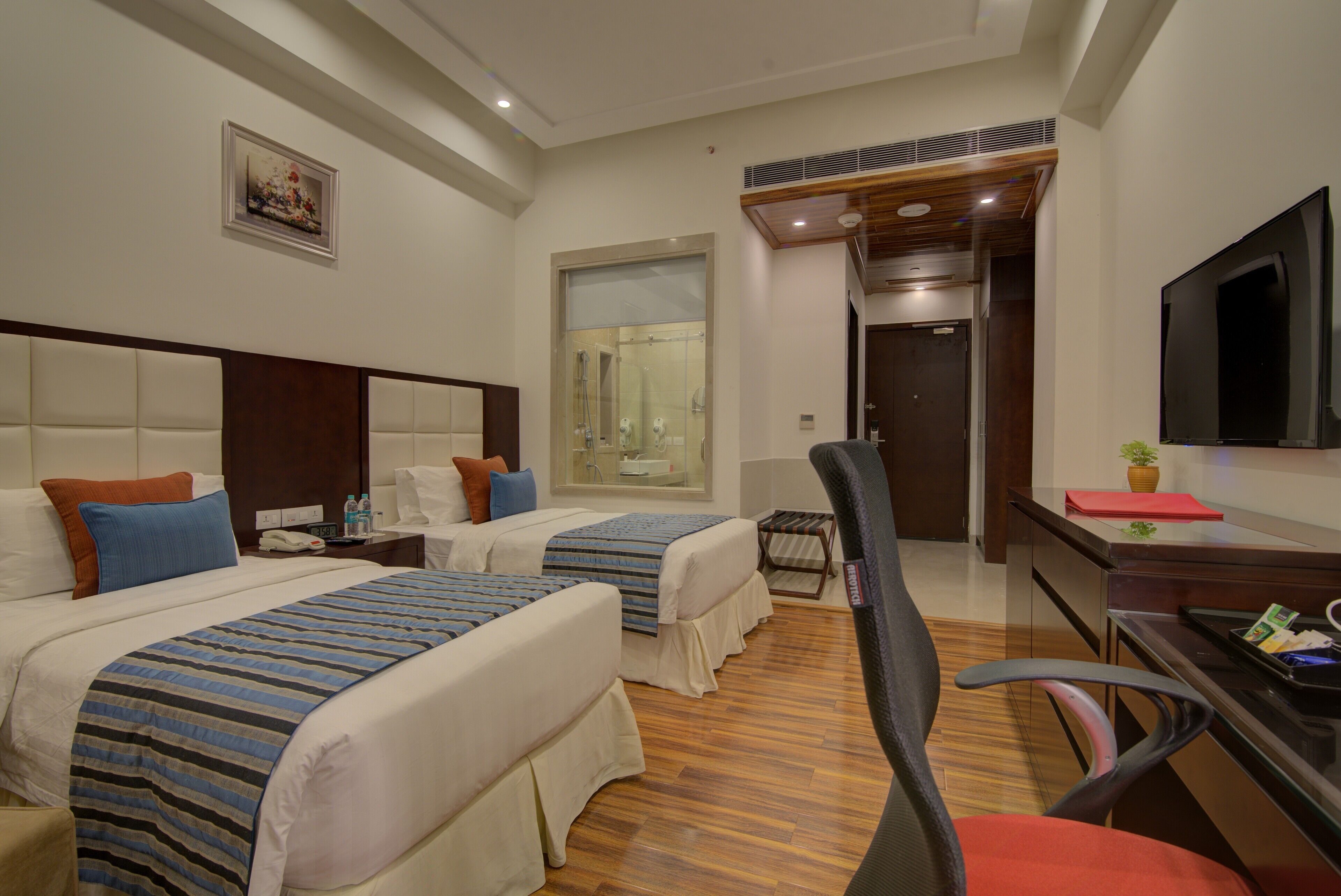 A hotel room with two beds, a desk, a chair, a TV, a shower area, and a painting on the wall.