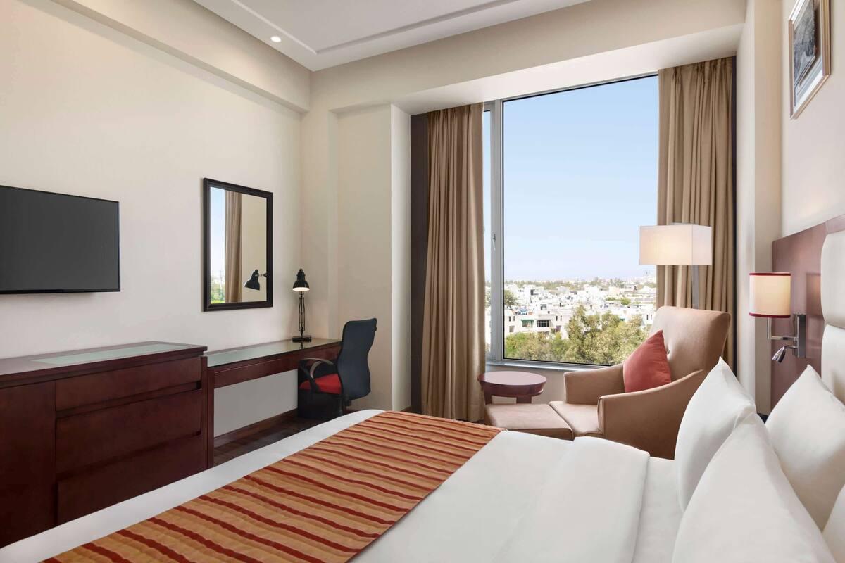 A hotel room with a large bed, a desk, a chair, a small table, and a view of the city through a large window.