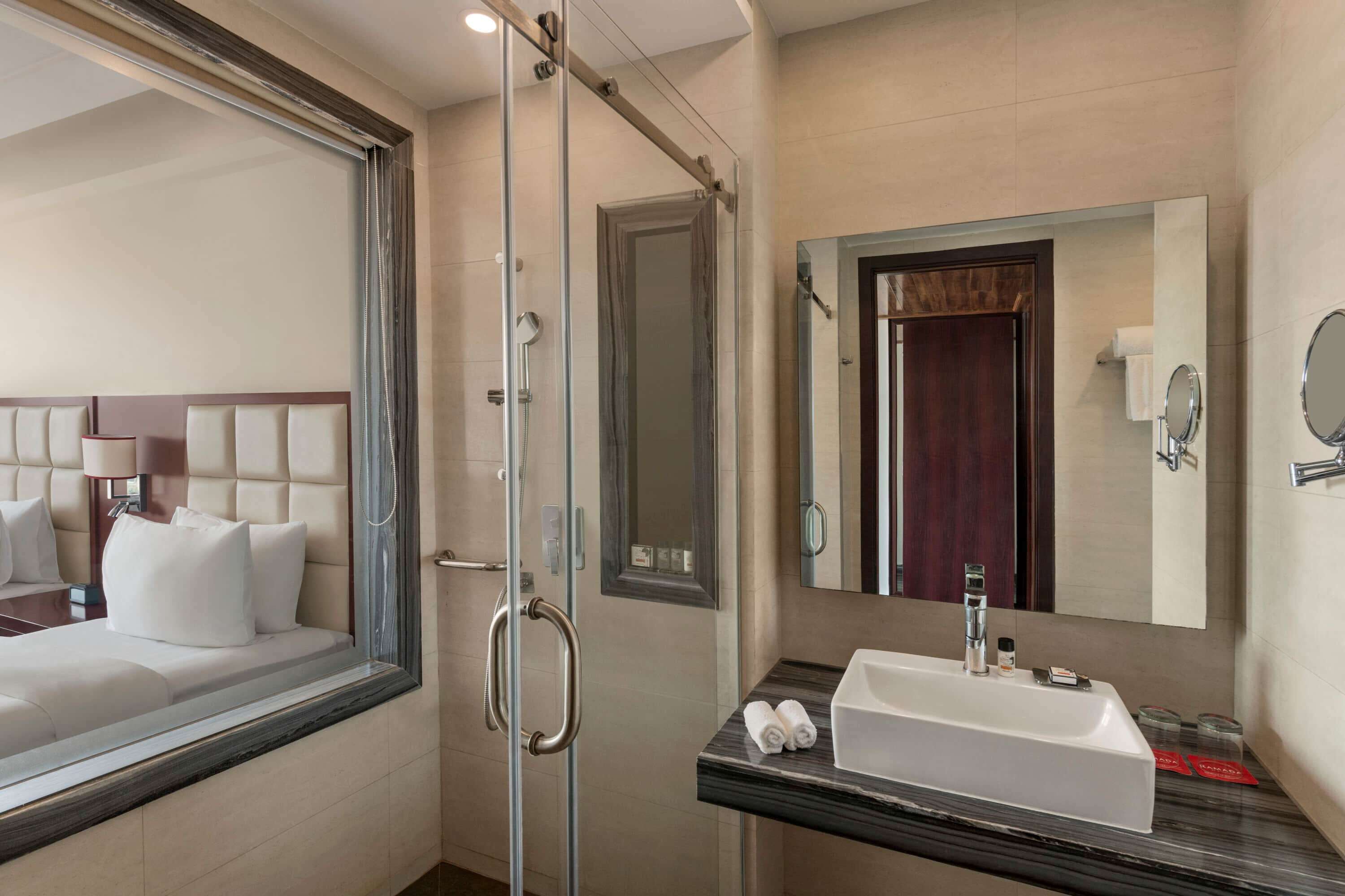 Ein Hotelbad mit einer gläsernen Dusche, einem großen Spiegel, einem Waschbecken und einem Bett mit Kopfteil.