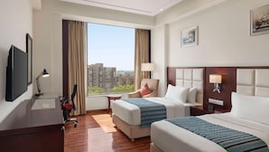 Habitación de hotel con dos camas, un escritorio, un televisor y un amplio ventanal con vista a la ciudad.