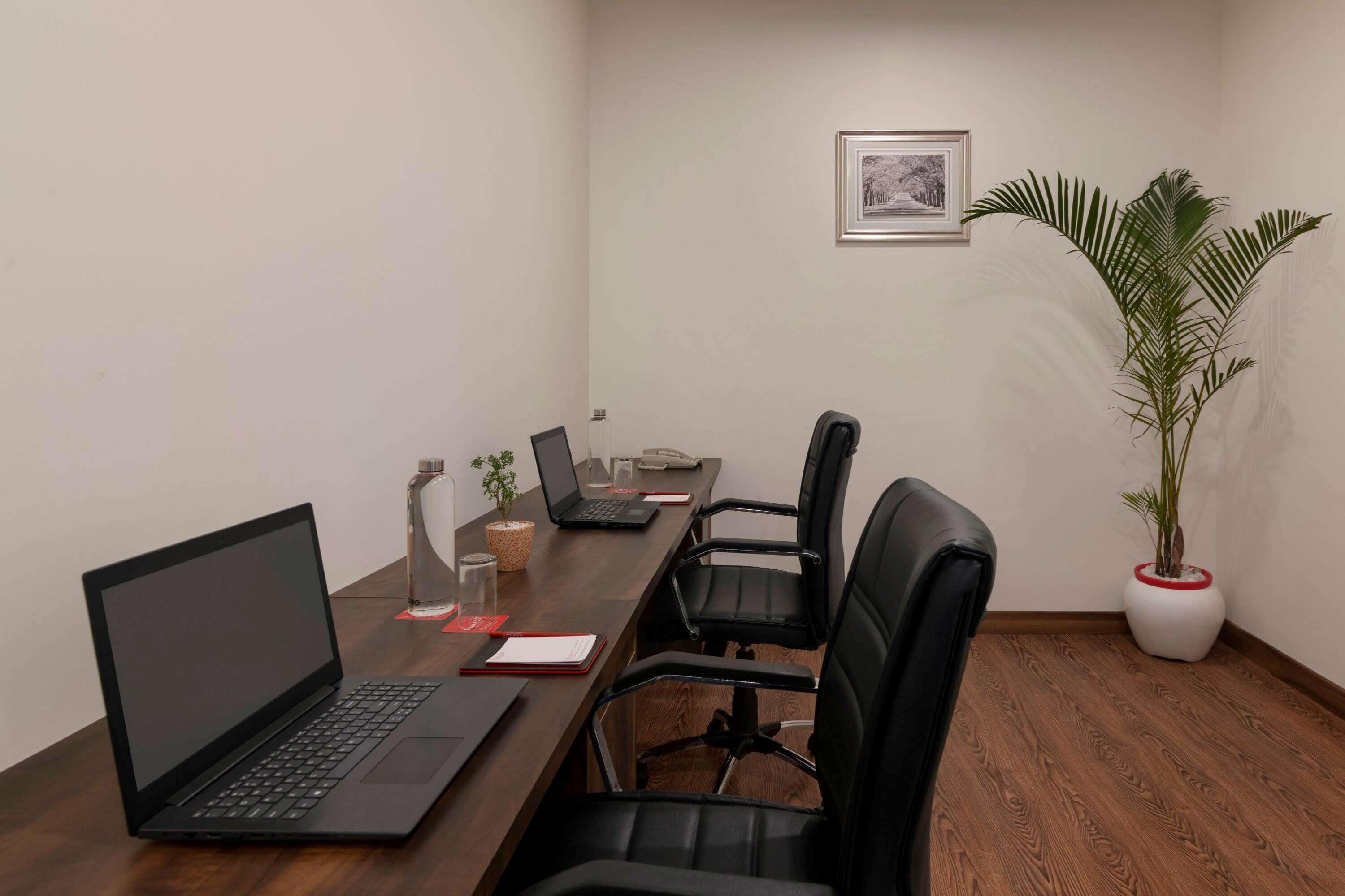 Sala de conferências com mesa de madeira, cadeiras pretas, um laptop, um vaso de plantas e um quadro pendurado na parede.