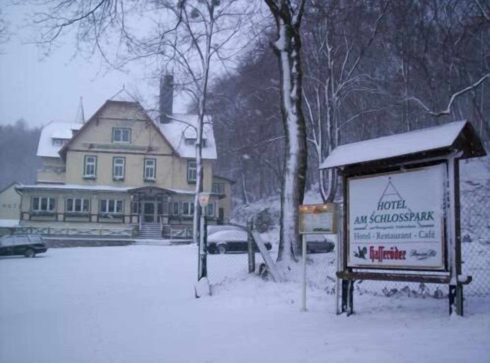 A snow-covered hotel sign with the name "Hotel Am Schlosspark" indicating it offers a hotel, restaurant, and café.
