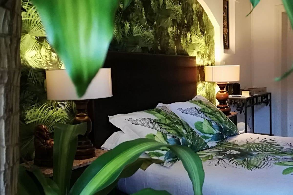 Un dormitorio con cama, dos mesitas de noche, una lámpara y una planta grande en maceta.