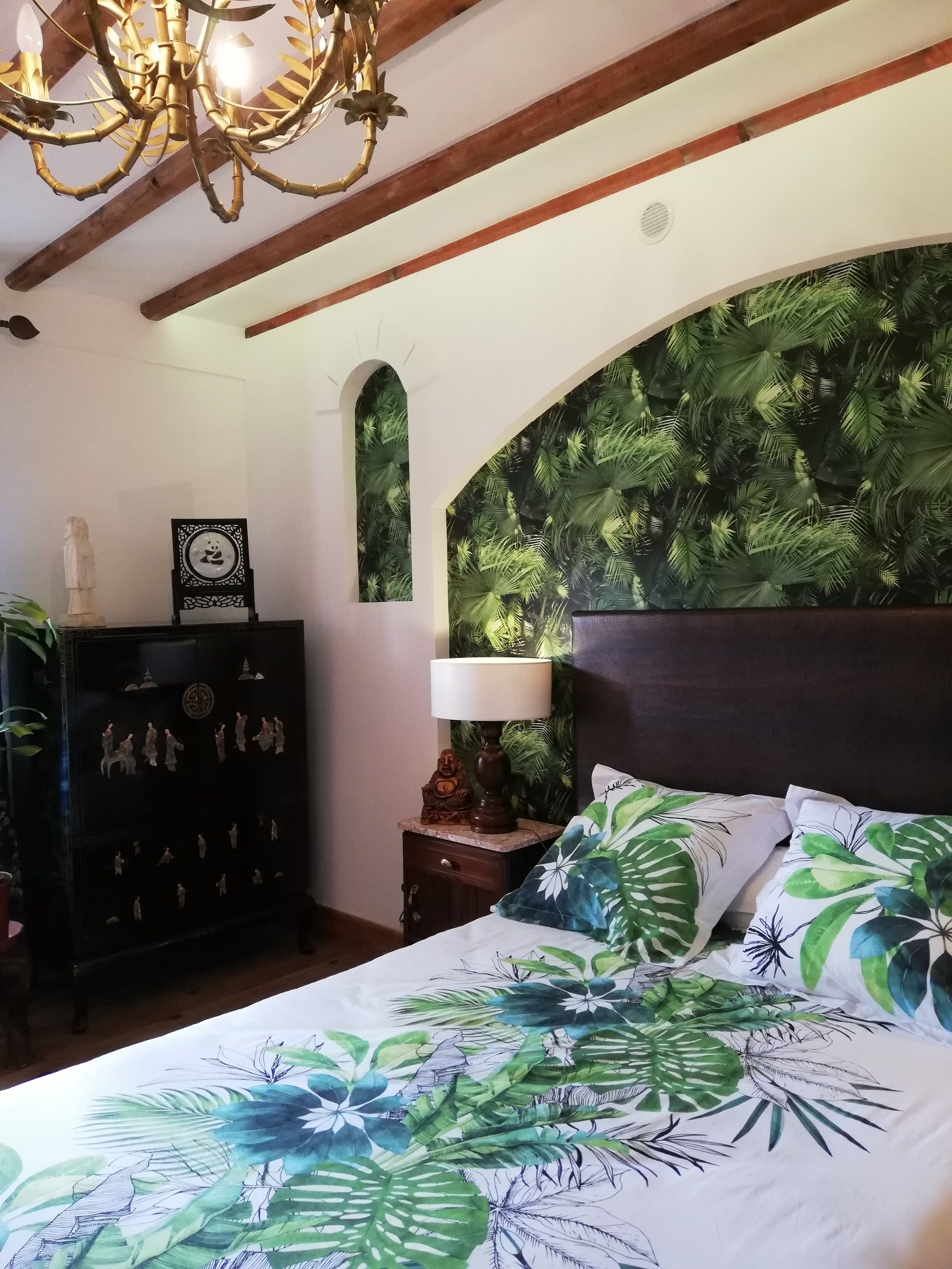 A bedroom with a bed, a nightstand, a wall-mounted shelf with decorative items, and a ceiling with exposed wooden beams and a chandelier.