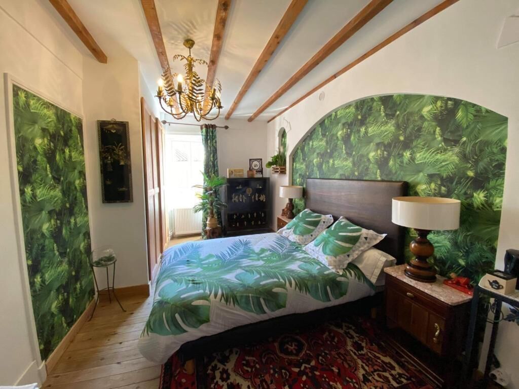 A bedroom with a bed, a nightstand, a lamp, a dresser, and a large plant.