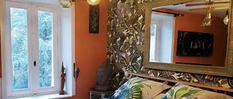 Un dormitorio con cama, ventana, espejo y pared con diseño de hojas tropicales.